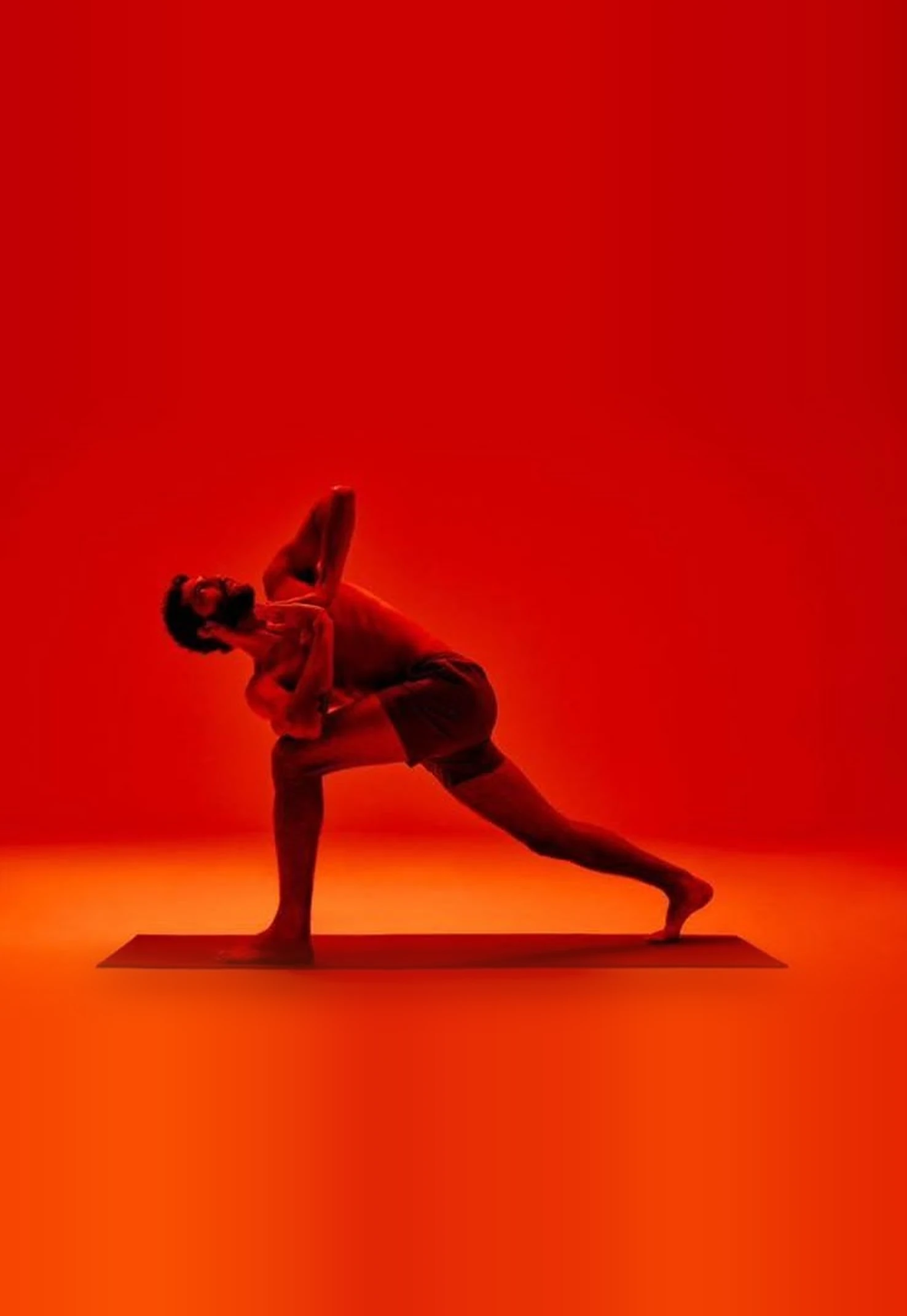 A man holding a deep twisted lunge on a dark mat, his silhouette lit by warm ambient red light